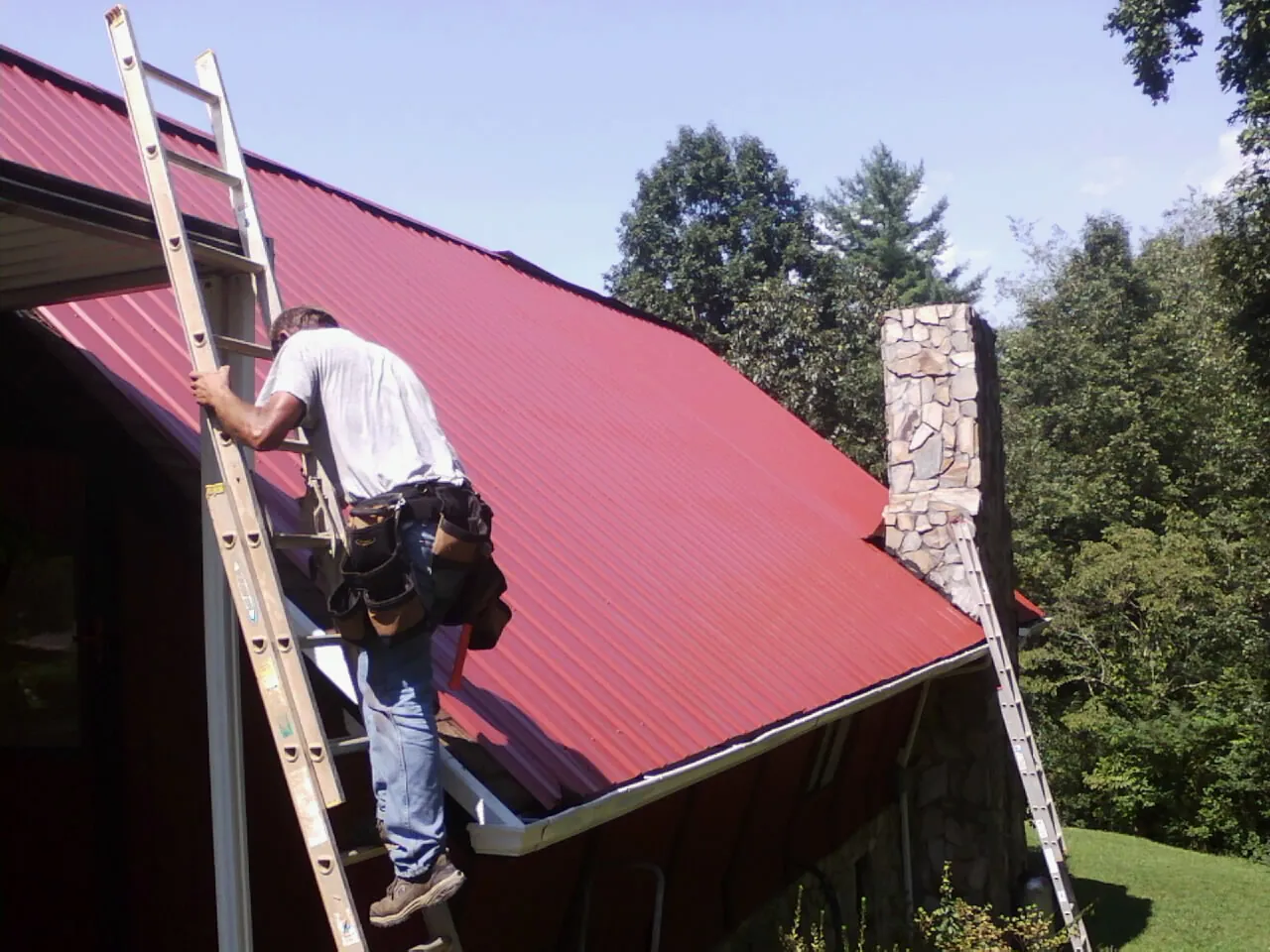 Roofer on a ladder beside a red metal roof and stone chimney — Soffit & Fascia Repair by Roofing Brothers in Delphi Falls