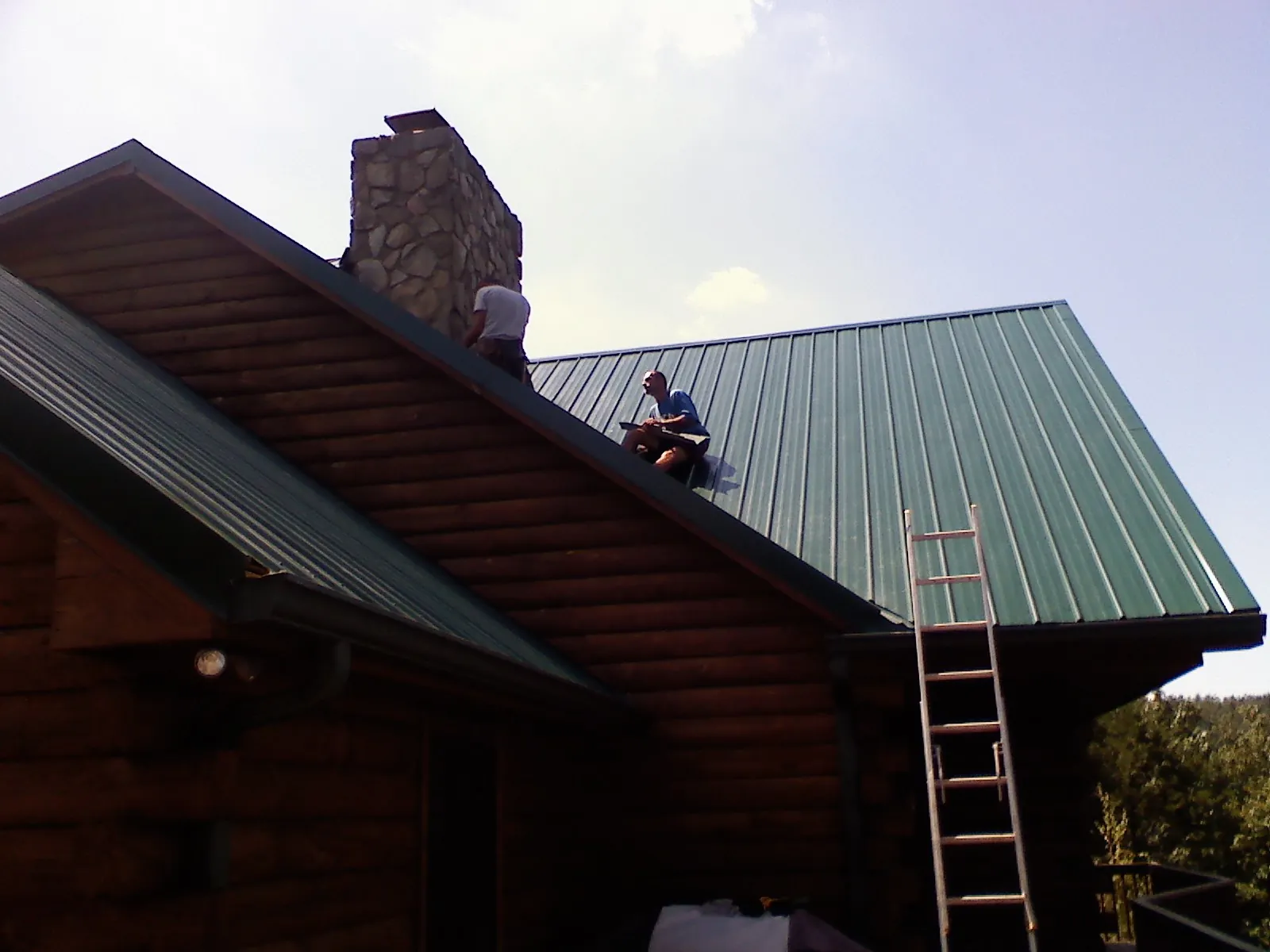 Roofers installing green metal panels beside a stone chimney on a log home in Delphi Falls